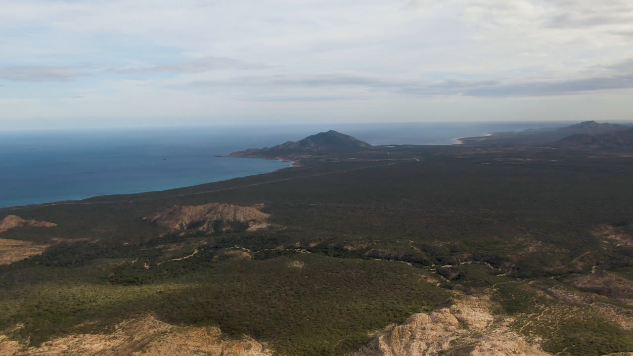 Wide aerial coastline view with low mountains, ocean, scrubland, and overcast sky.