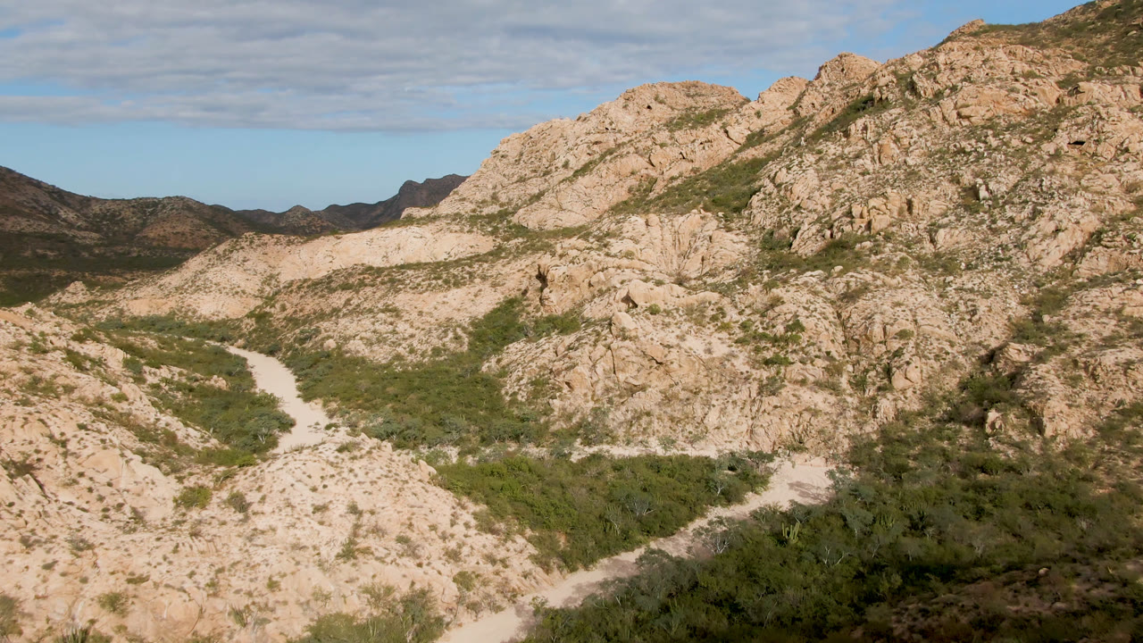 Elevated Cabo Pulmo mountain view with tan rock ridges, green scrub in the valley floor, a pale curved route at left, and no visible people.
