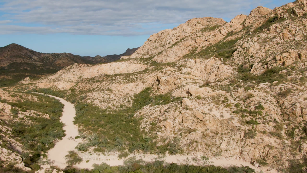 Wide view of a pale dirt road curving through rocky desert terrain in Cabo Pulmo, Baja California Sur, with green scrub, tan hills, distant dark ridges, and no visible people.