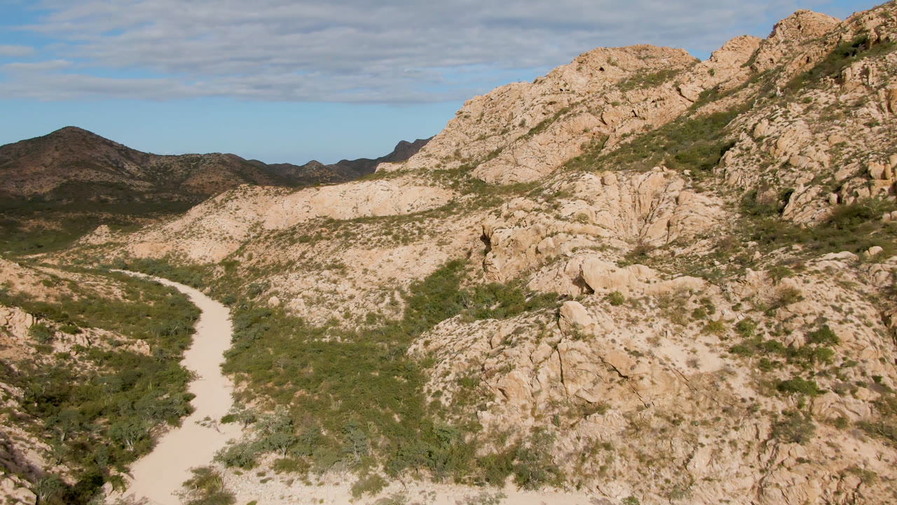 Elevated landscape frame in Cabo Pulmo, Baja California Sur, with the dirt road bending through low green vegetation below tan rock formations and no visible people.