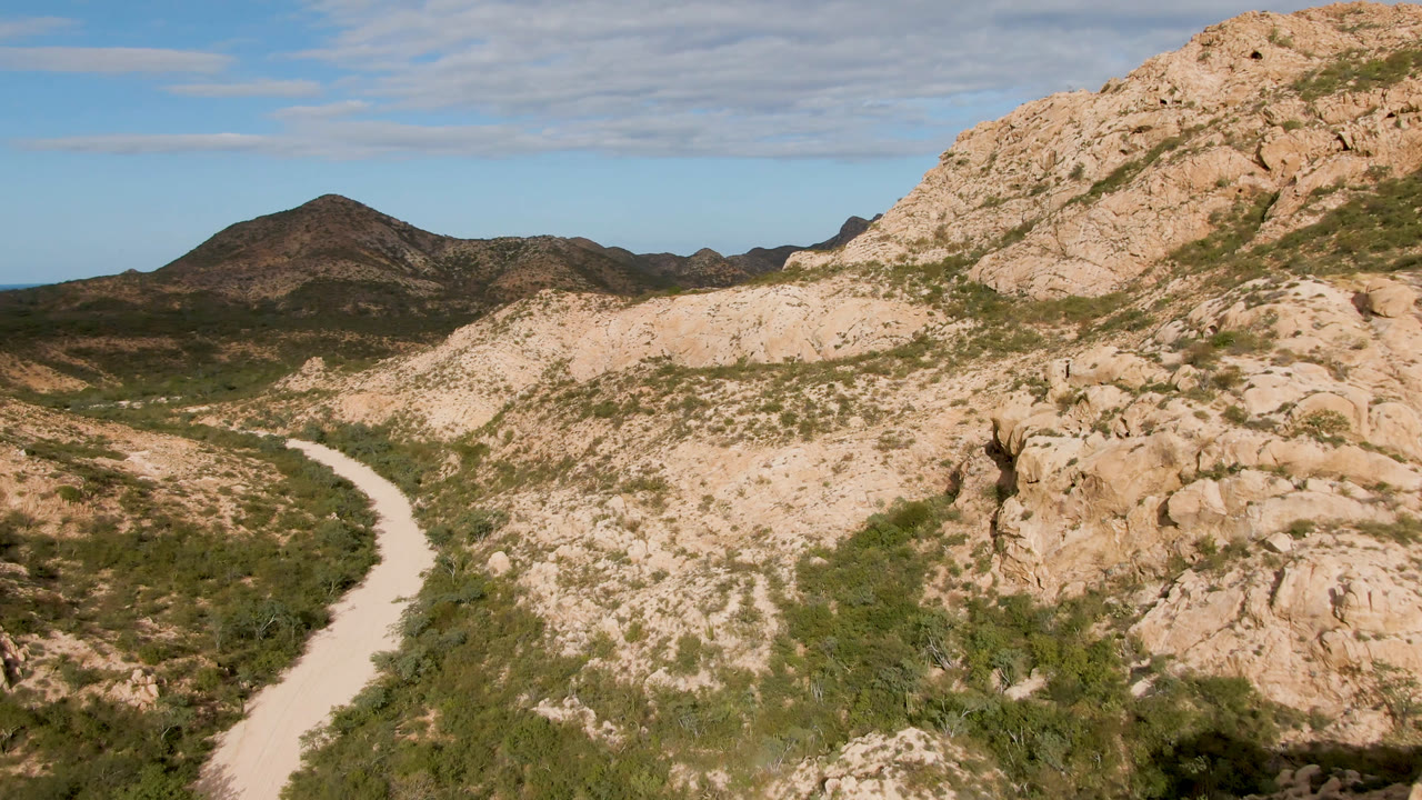 Wide view in Cabo Pulmo, Baja California Sur, showing the road sweeping up from the lower left toward scrub-covered slopes, tan rock faces, distant hills, and no visible people.