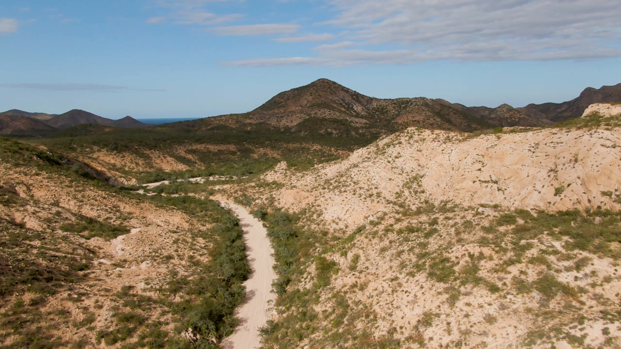 Elevated view in Cabo Pulmo, Baja California Sur, Mexico, showing a pale dirt road curving through scrubby desert hills toward dark mountains, with no visible people.