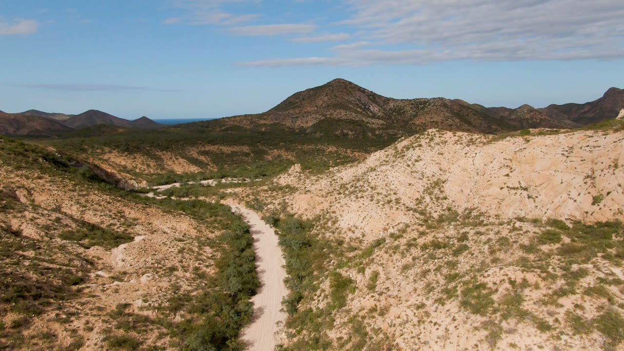 Cabo Pulmo aerial-style frame with the pale road tighter along the lower left valley, tan hillside in front, distant mountain ridges, blue sky, and no visible people.