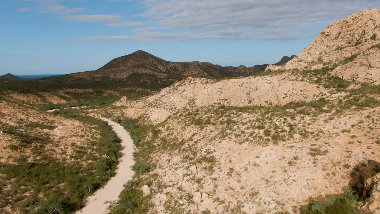 At 0:00, a pale dirt road curves through scrub-covered hills in Cabo Pulmo, Baja California Sur, Mexico, with low mountains and no visible people.