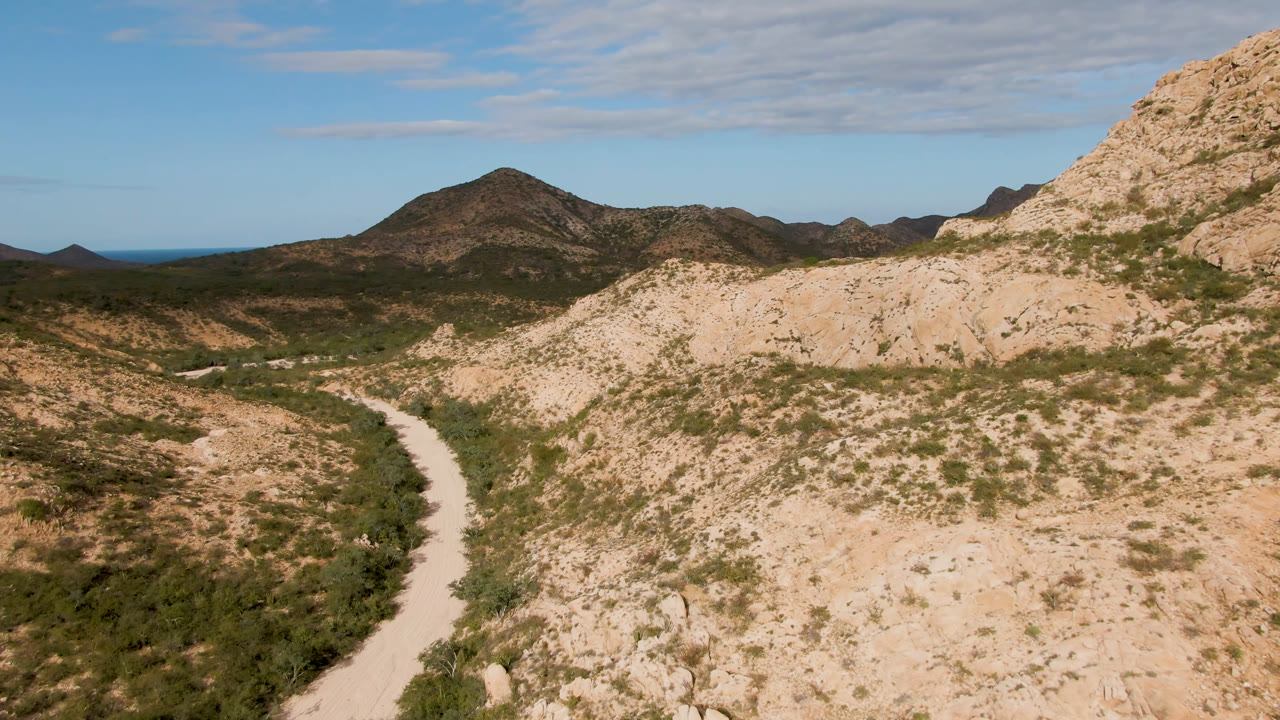 At 0:01, the dirt road sits slightly left of center as tan rocky slopes and green brush lead toward distant mountains in Cabo Pulmo, with no visible people.