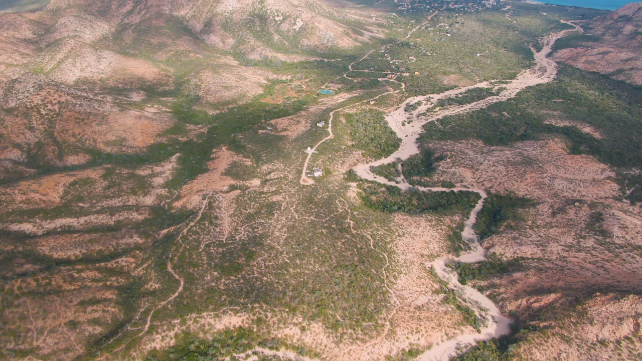 Aerial frame over Cabo Pulmo, Baja California Sur, showing a pale road and dry wash cutting through green-brown mountain terrain with no visible people.
