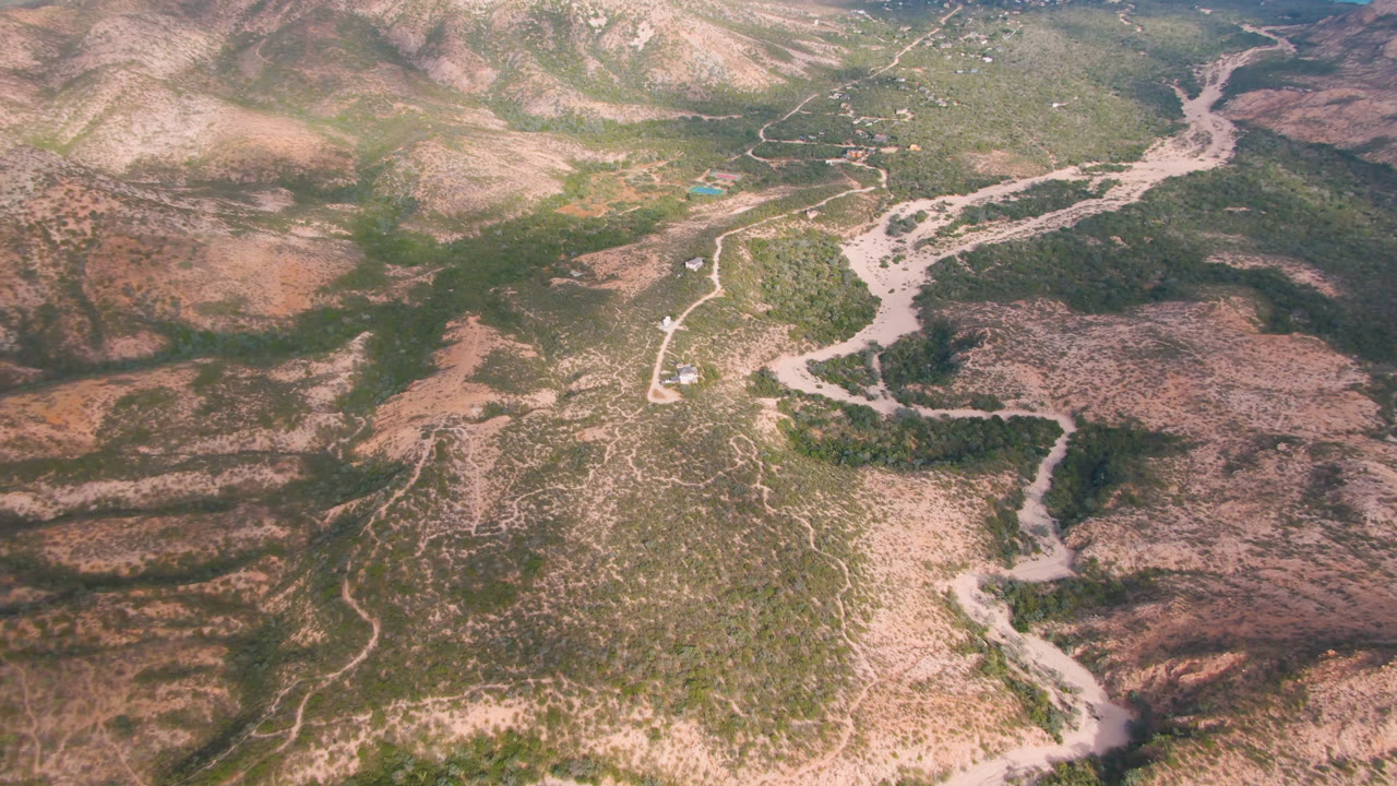 A second aerial moment over Cabo Pulmo, Baja California Sur, with the dirt road lower in frame and branching dry river channels across the valley, no visible people.