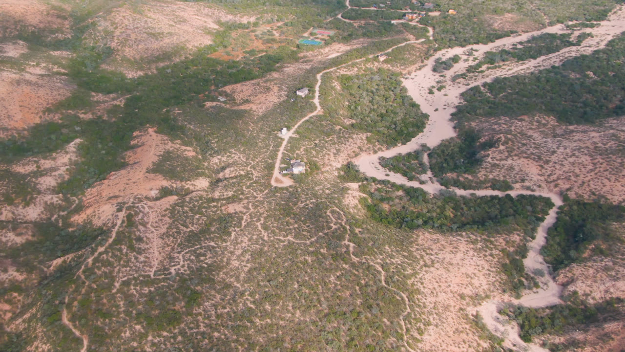 Aerial frame over Cabo Pulmo, Baja California Sur, showing a pale winding dirt road through dry scrubland, sandy wash lines, low vegetation, and no visible people.