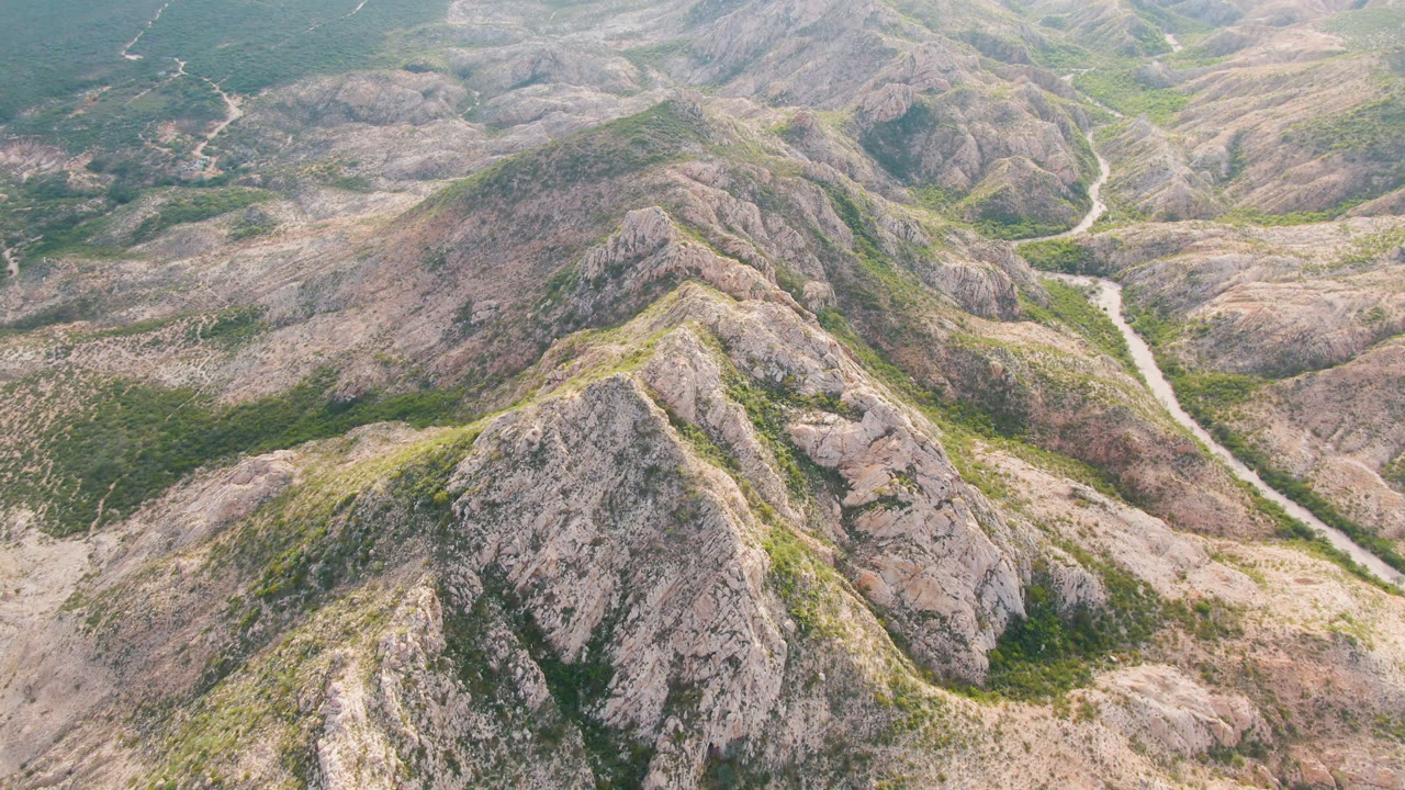 Aerial frame over Cabo Pulmo ridges where gray-tan slopes surround a narrow dirt road through the middle of the landscape, with no visible people.