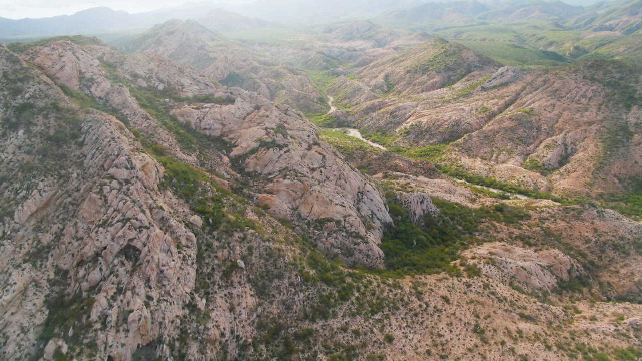 Wide aerial frame over Cabo Pulmo rocky ridges with green scrub in the valley floor, hazy mountains in the distance, and no visible people.