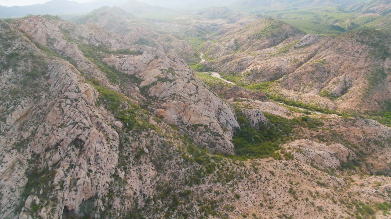 Aerial view in Cabo Pulmo looking across a steep gray-tan ridge toward a winding pale road or wash through green valley vegetation, with no visible people.