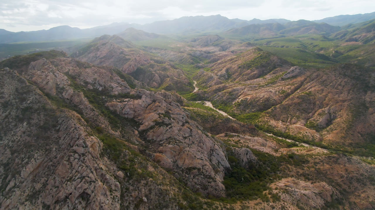 Aerial view over rocky hills near Cabo Pulmo, Baja California Sur, with a winding dirt road crossing the green valley floor and no visible people.