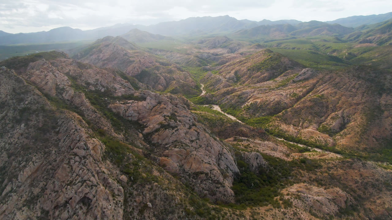 High aerial frame of Cabo Pulmo mountain terrain, showing pale rock faces, scrub vegetation, a road curving through the center valley, and no visible people.