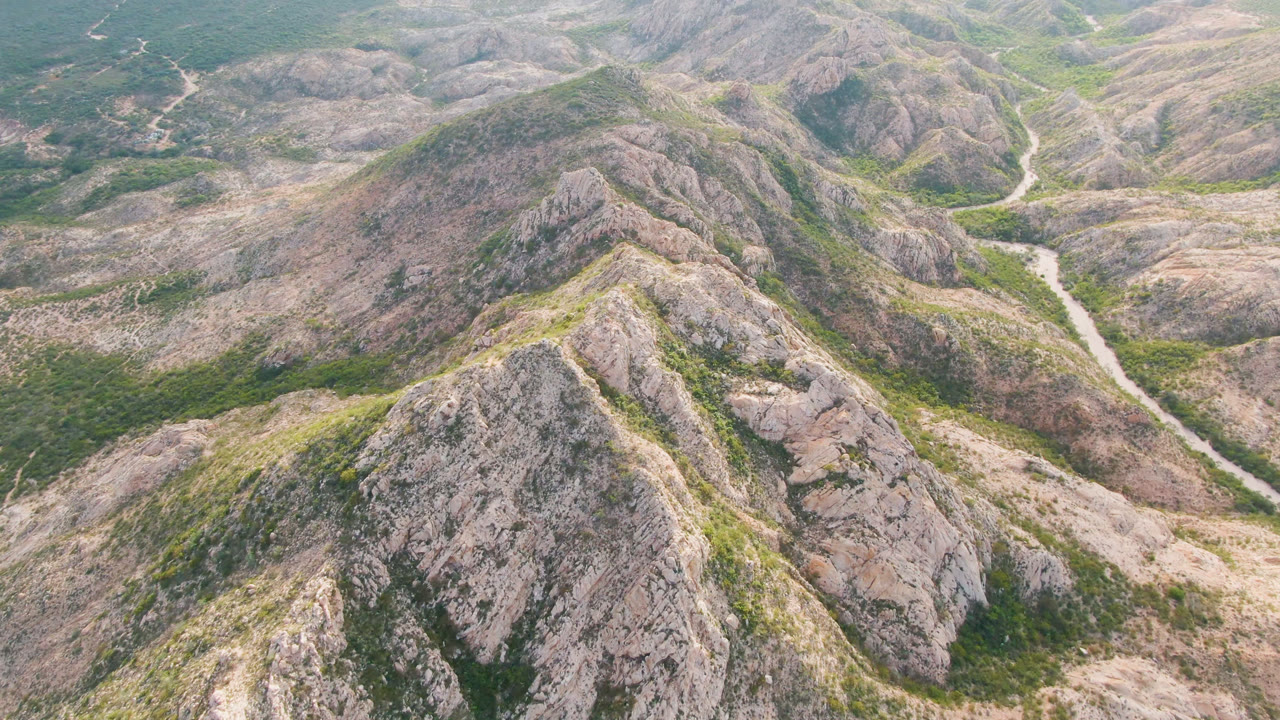Aerial view over rocky Cabo Pulmo hills with green patches, tan ridges, a pale winding road or wash in the valley, and no visible people.