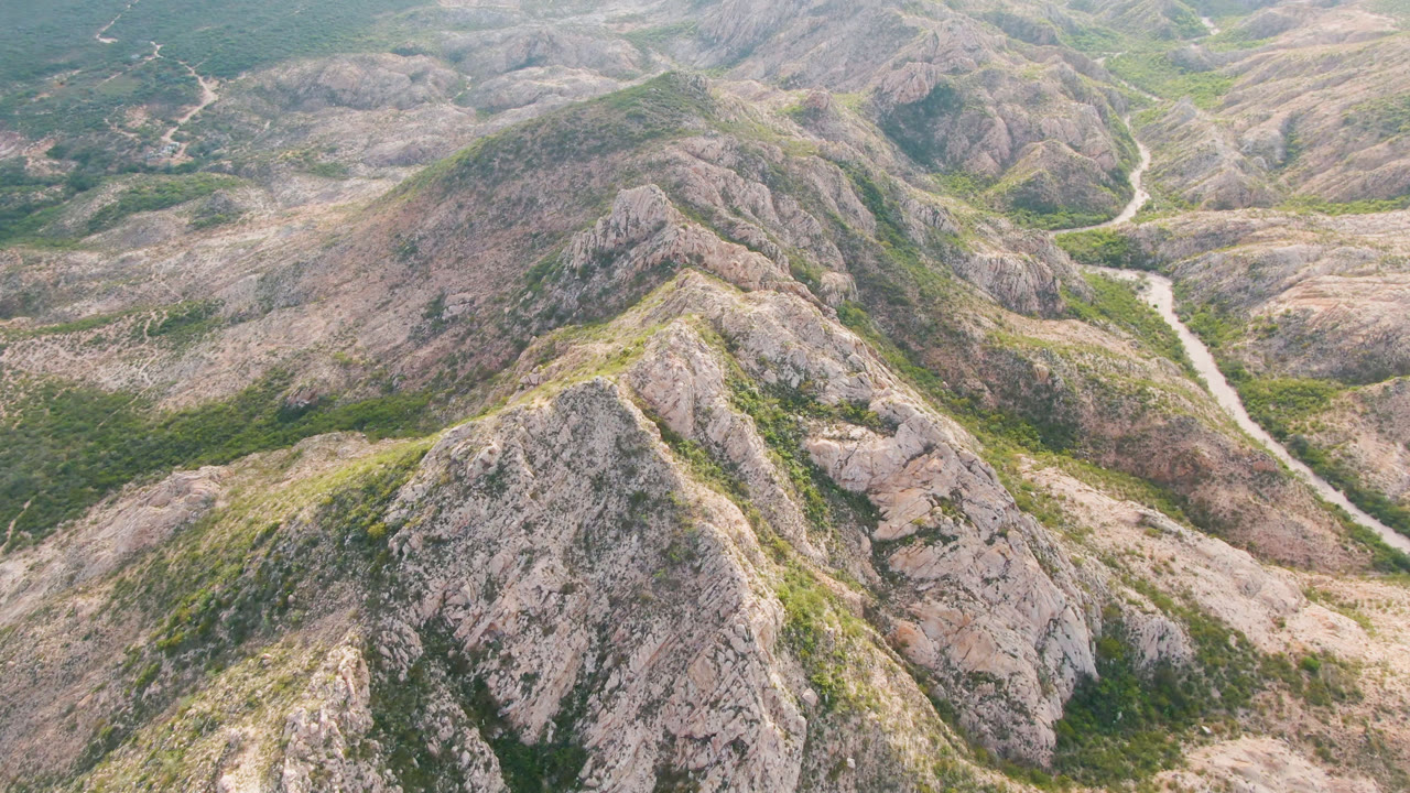 High aerial frame of Cabo Pulmo's rocky terrain, with a central ridge in front, a pale valley track to the right, sparse green vegetation, and no visible people.