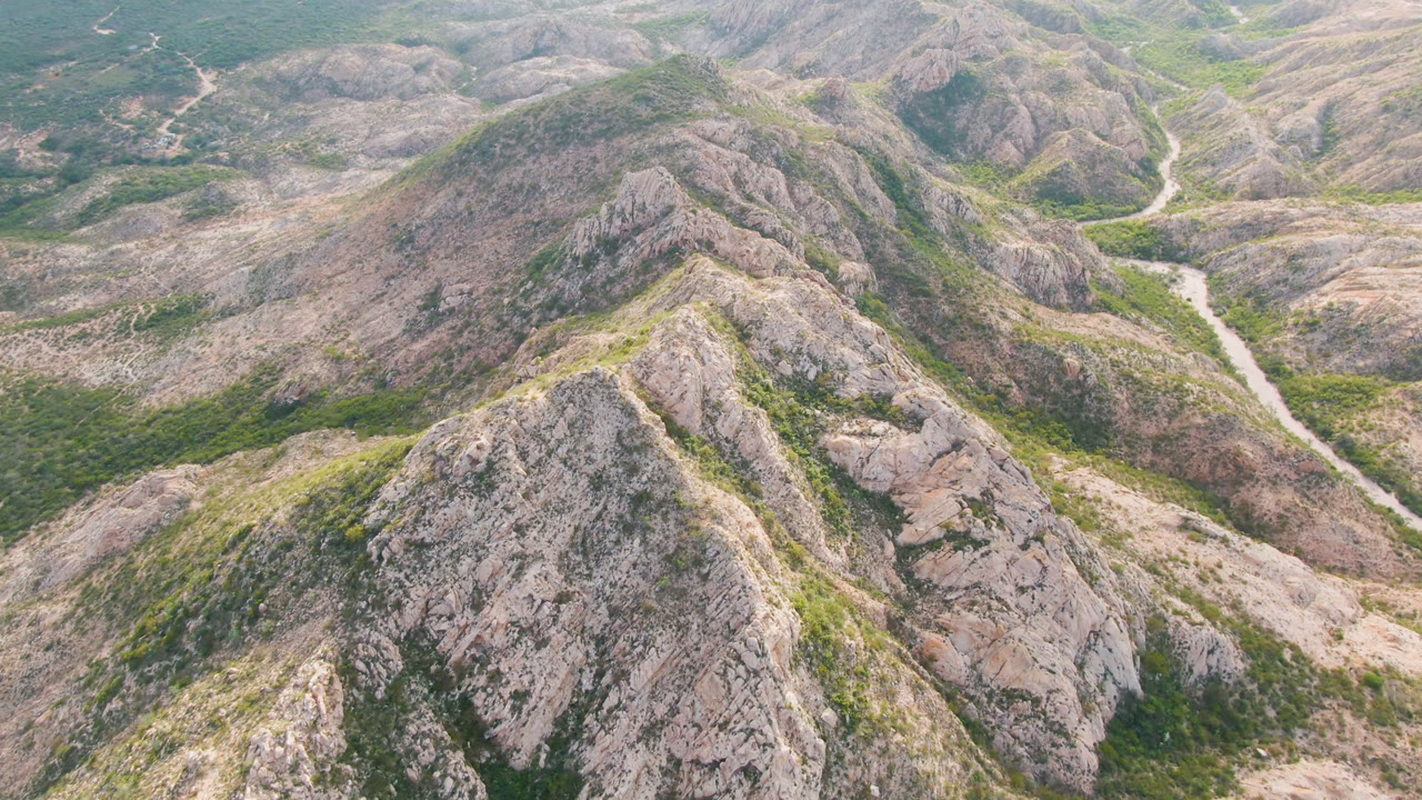 Opening aerial frame over Cabo Pulmo rocky hills, with pale ridges, green scrub, a winding dirt road in the middle distance, and no visible people.