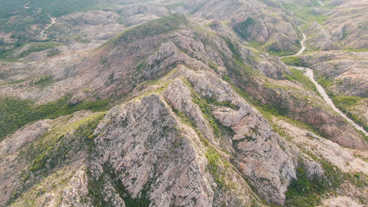 Aerial frame over Cabo Pulmo showing a central rock ridge, patchy green vegetation, a pale winding road beyond it, and no visible people.