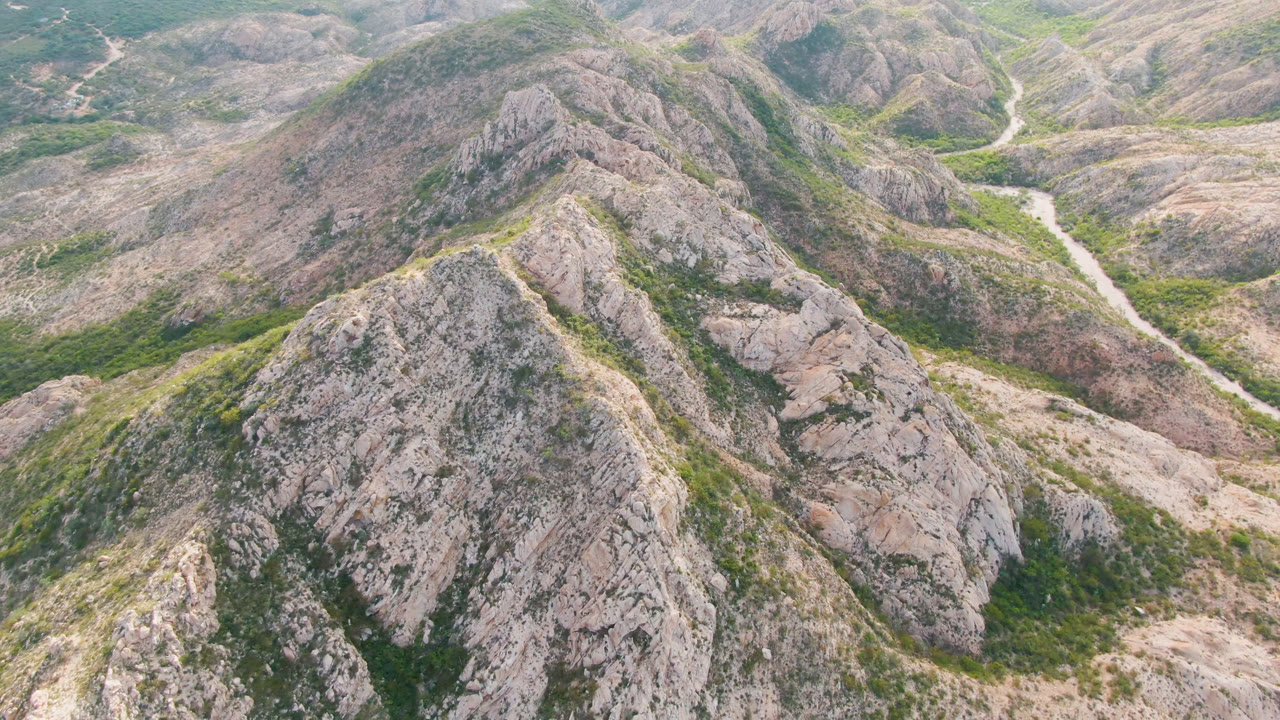 Aerial frame over Cabo Pulmo rocky hills, with green scrub on pale ridges, a light dirt road bending through the valley, and no visible people.