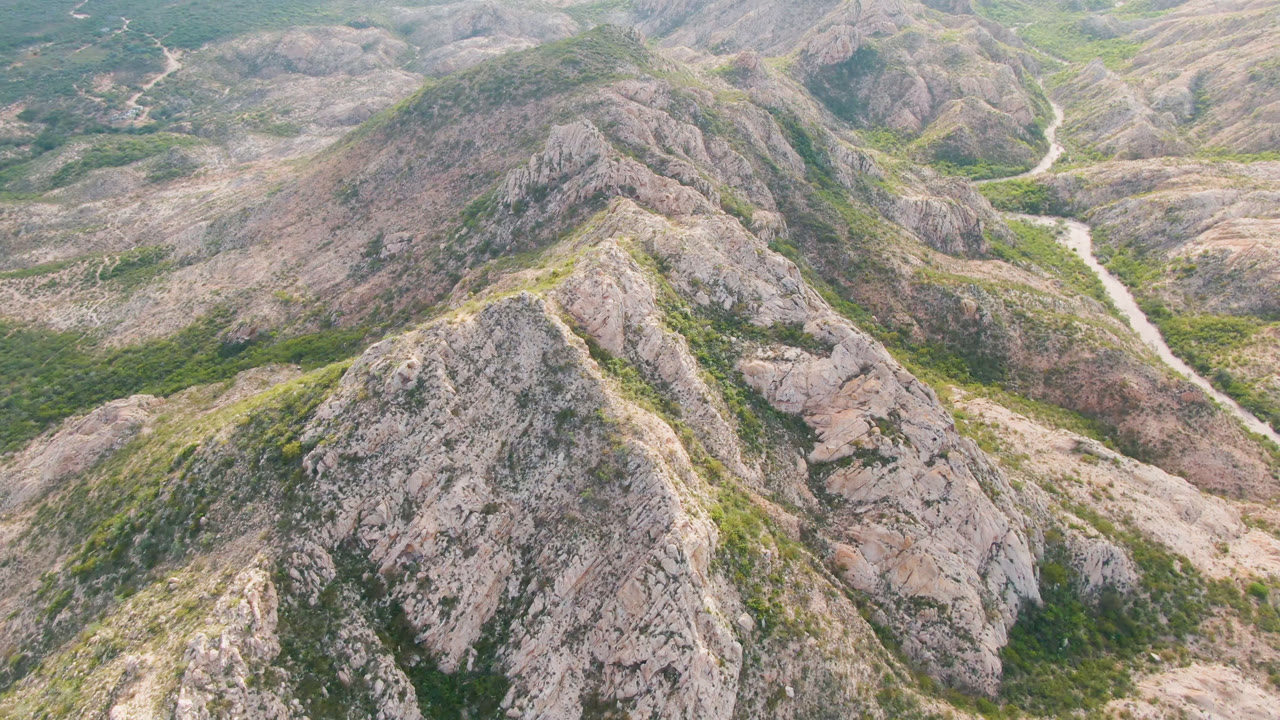 Aerial frame over Cabo Pulmo, Baja California Sur, Mexico, showing rocky ridges, green scrub in the gullies, and a pale winding road or wash near the center with no visible people.