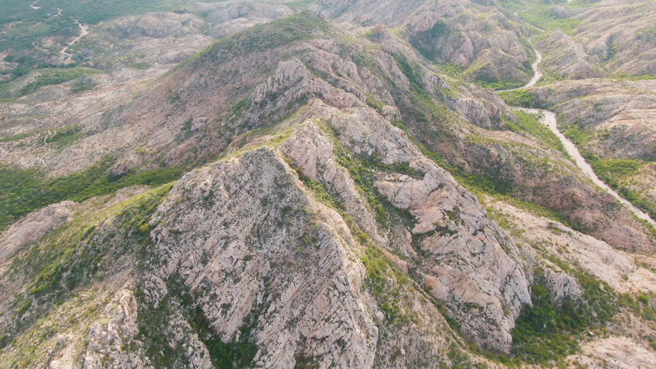 Aerial view in Cabo Pulmo, Baja California Sur, Mexico, with a central gray-tan ridge, sparse green vegetation, and a pale winding track or wash cutting through the valley, with no visible people.