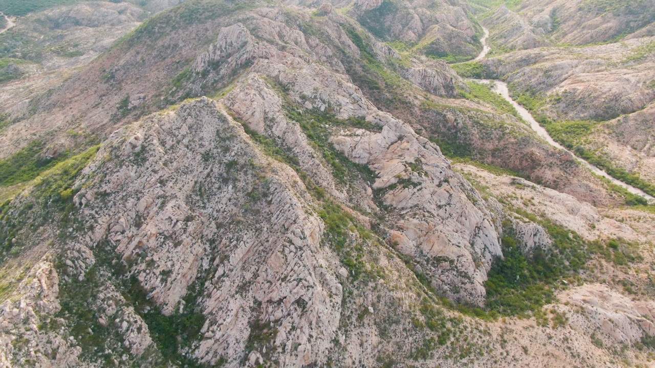 Aerial view in Cabo Pulmo with a central rocky ridge in front, scattered green vegetation on slopes, and a pale winding channel visible in the valley behind, with no visible people.
