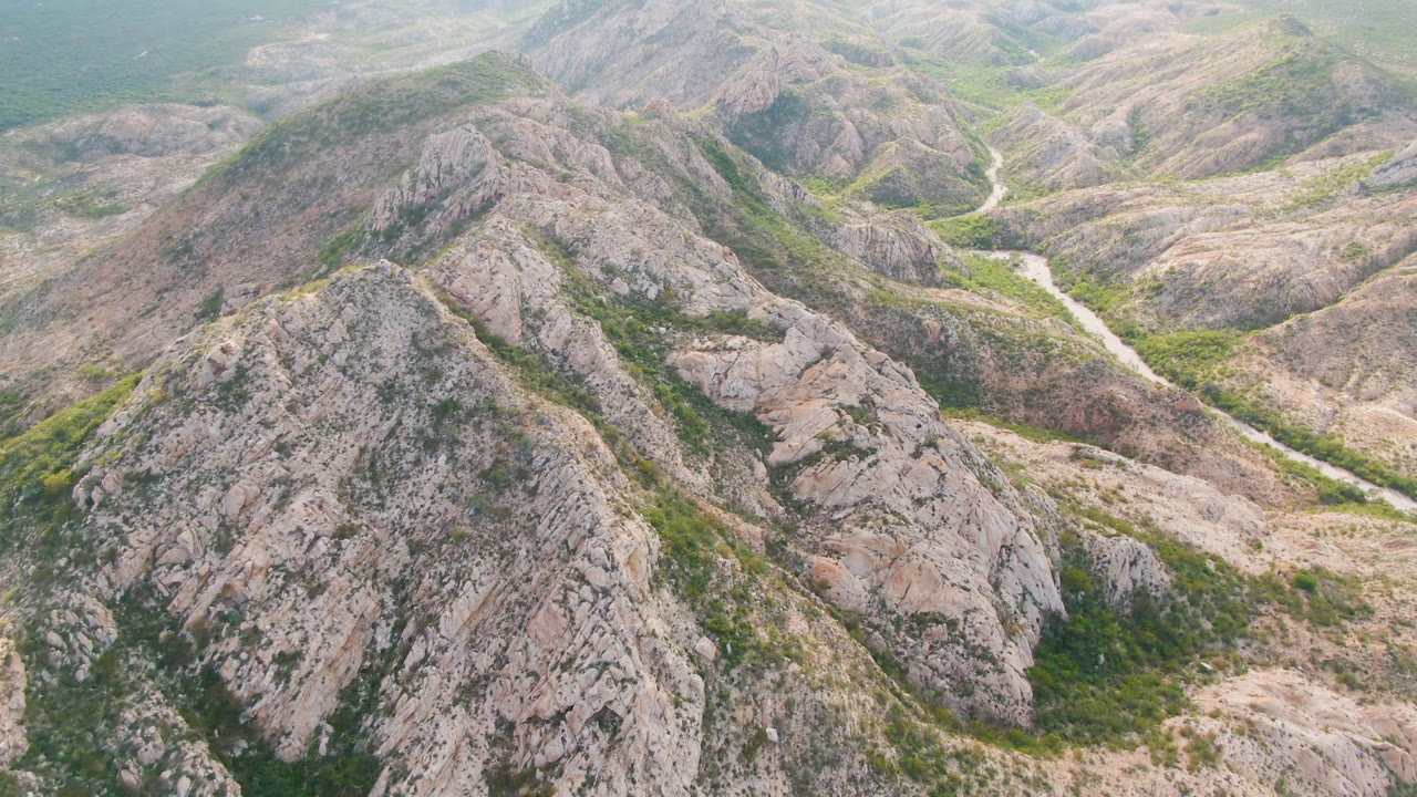 Aerial frame from Cabo Pulmo, Baja California Sur, showing rocky tan ridges, green gullies, a pale river corridor in the valley, and no visible people.