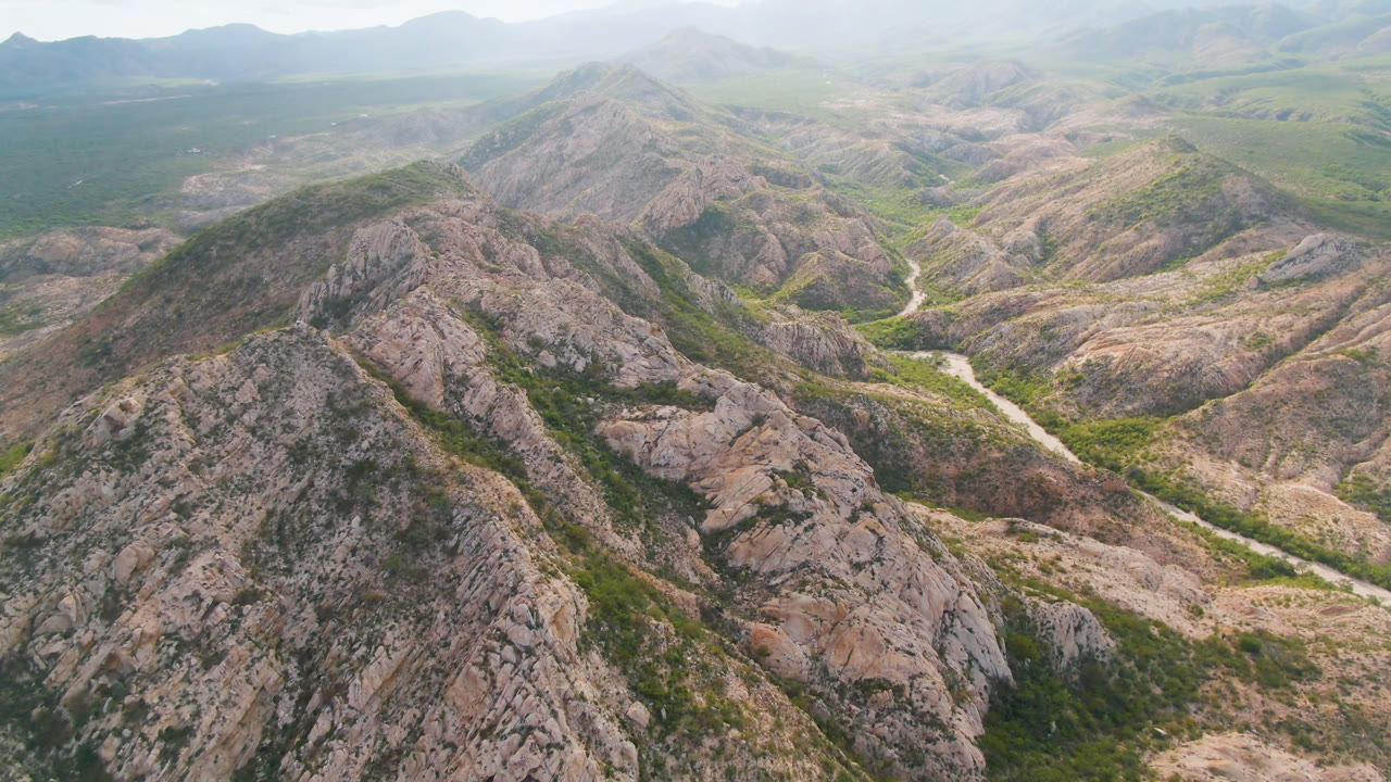 Aerial frame over Cabo Pulmo, Baja California Sur, showing a rocky foreground ridge, green valley floor, a pale winding river or wash, distant mountains, and no visible people.
