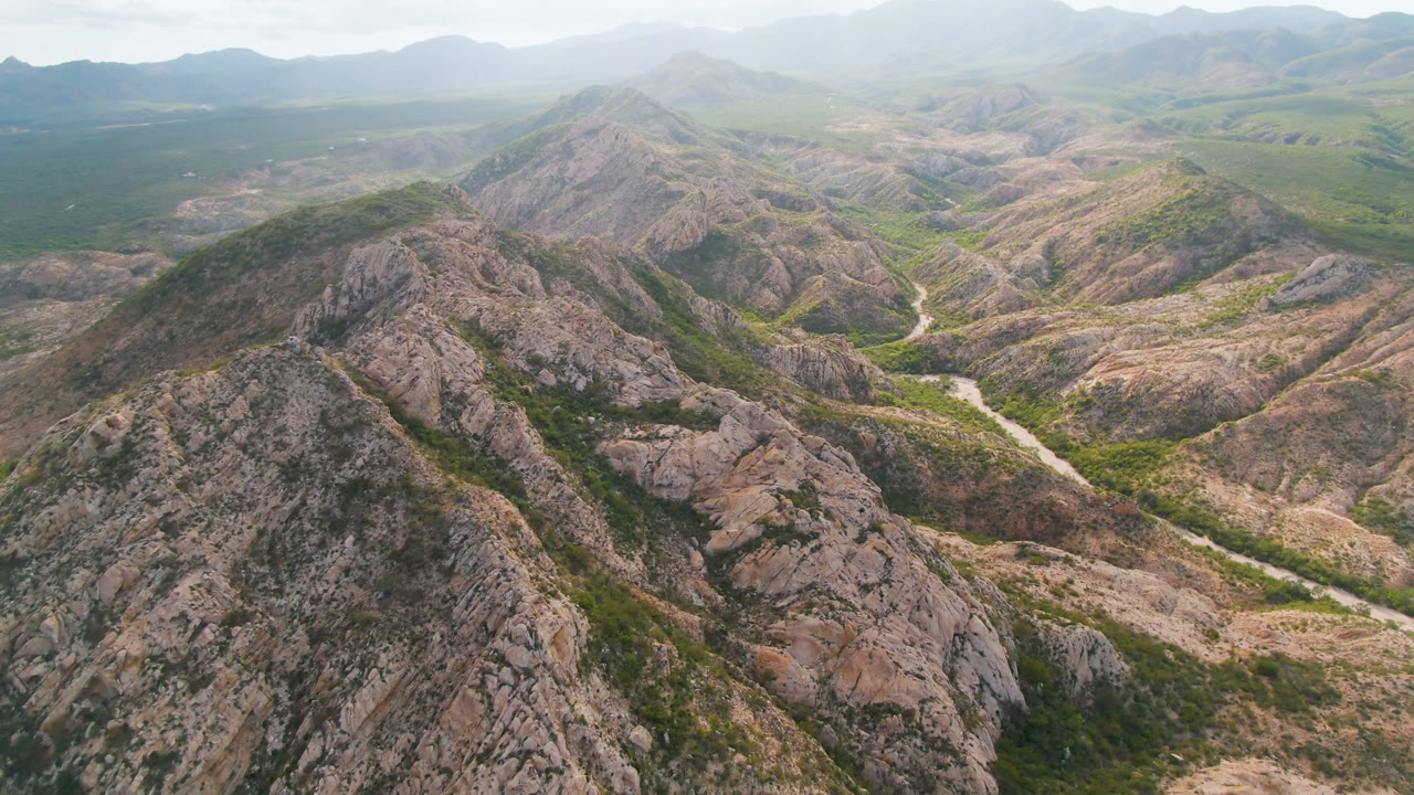 Second aerial frame from Cabo Pulmo, Baja California Sur, with the central rocky ridge filling the lower frame, a narrow light-colored watercourse through the valley, hazy mountain layers, and no visible people.