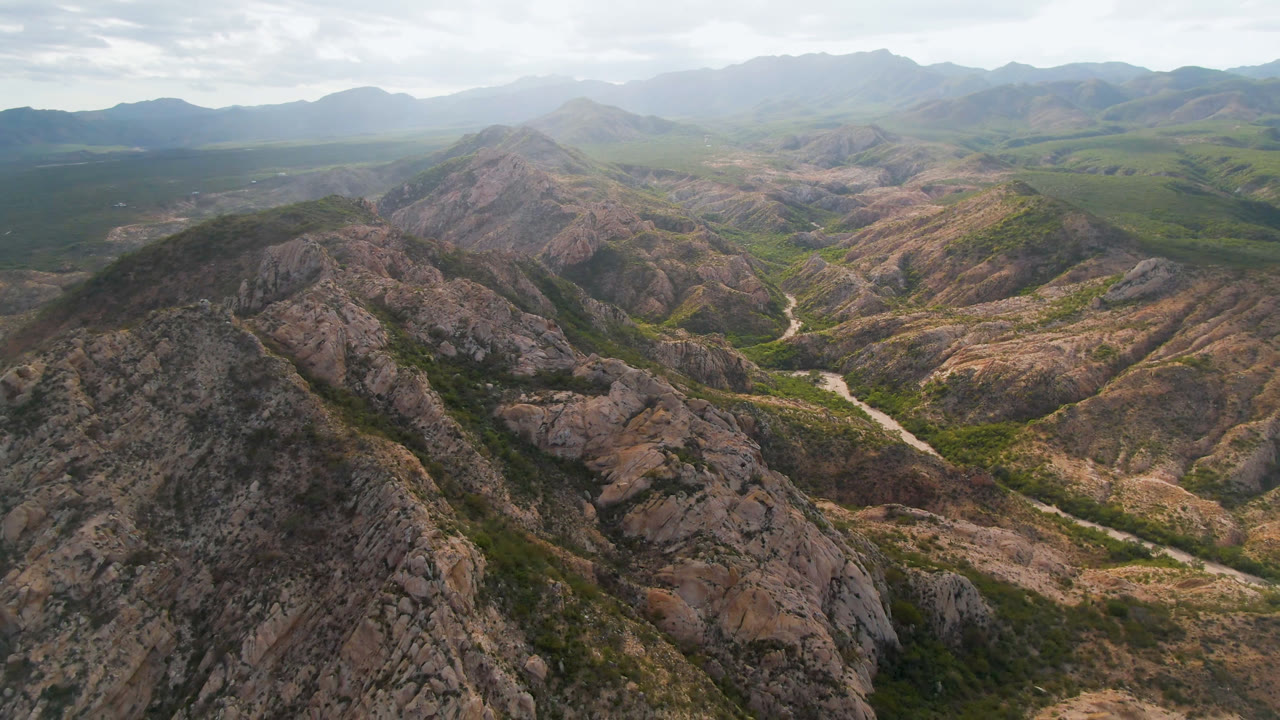 Aerial view over Cabo Pulmo rocky desert ridges with a pale river or wash curving through the valley, green vegetation in low areas, hazy mountains in the distance, and no visible people.
