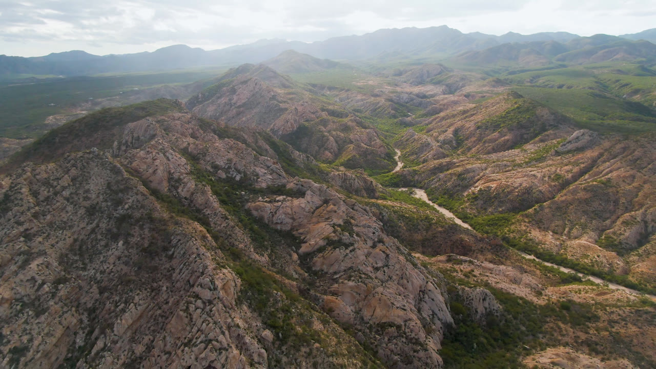 Wide aerial frame of Cabo Pulmo with a dark rocky ridge in the foreground, a winding pale river or wash in the center valley, green patches along the low ground, distant mountains, cloudy sky, and no visible people.