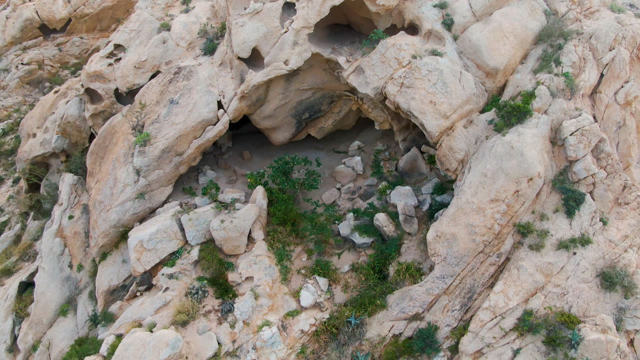 Overhead frame in Cabo Pulmo, Baja California Sur, Mexico showing pale eroded rock, a small dark cave recess near the upper left, scattered boulders, green shrubs, and no visible people.