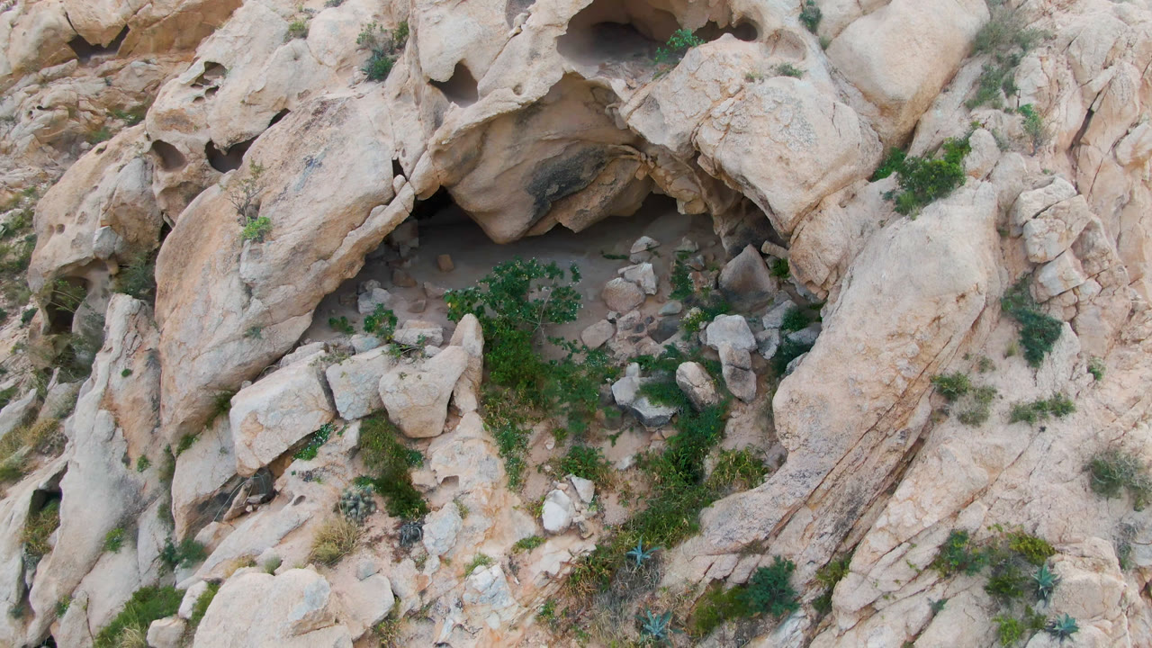 Second overhead frame in Cabo Pulmo shows the cave recess higher in the composition, loose stones below it, tan rock ridges, patches of green scrub, and no visible people.