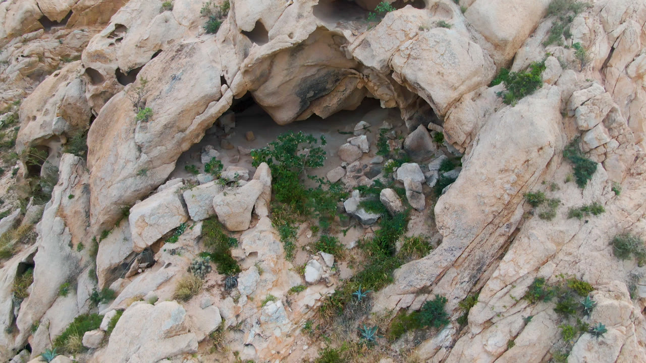 Aerial view of eroded tan stone around a cave with shrubs and broken rocks.