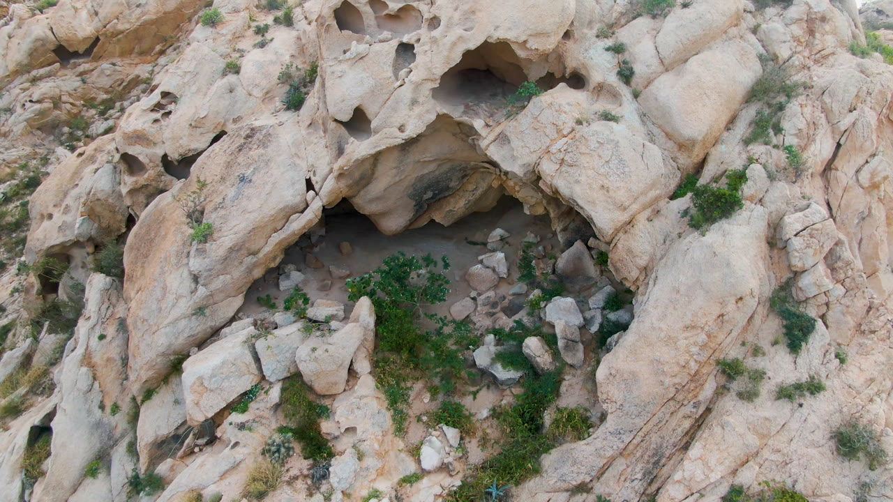 Overhead frame from Cabo Pulmo, Baja California Sur, Mexico showing a small cave opening in pale eroded rock, scattered boulders, green shrubs, and no visible people.