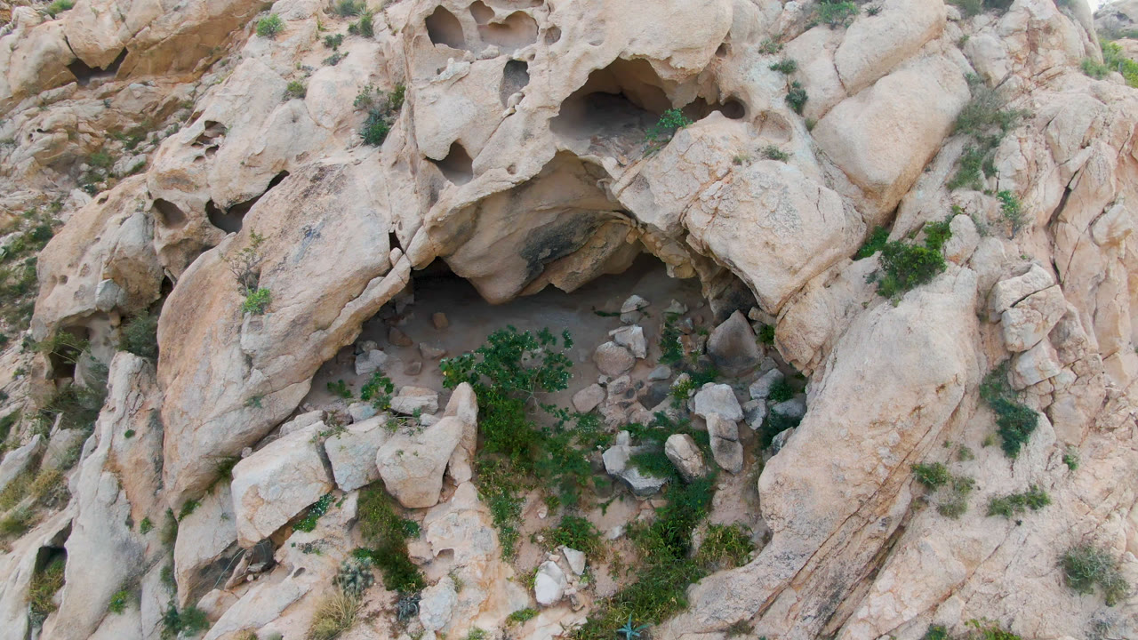 Aerial frame from Cabo Pulmo, Baja California Sur, Mexico with the cave recess near the upper center, tan rock ridges, loose stones, patches of scrub, and no visible people.