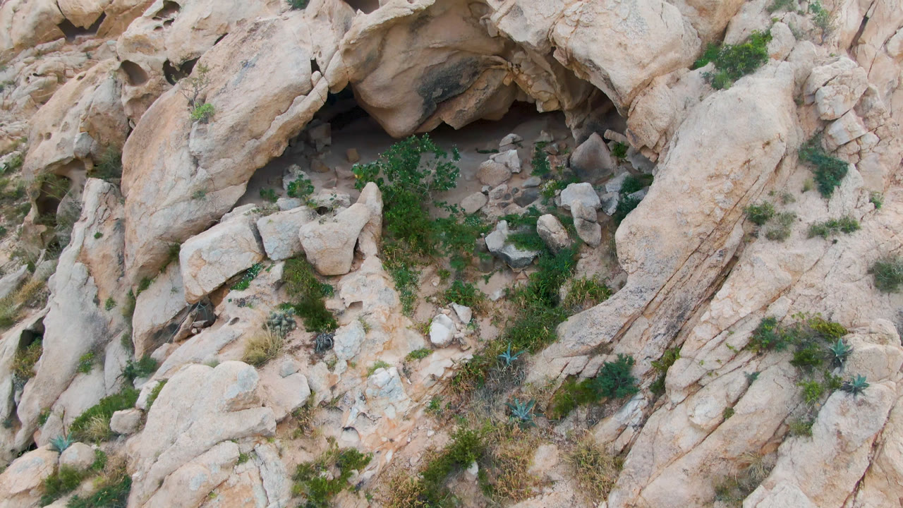 Overhead view in Cabo Pulmo, Baja California Sur, Mexico, showing pale rock, a dark cave opening, scattered boulders, sparse green growth, and no visible people.