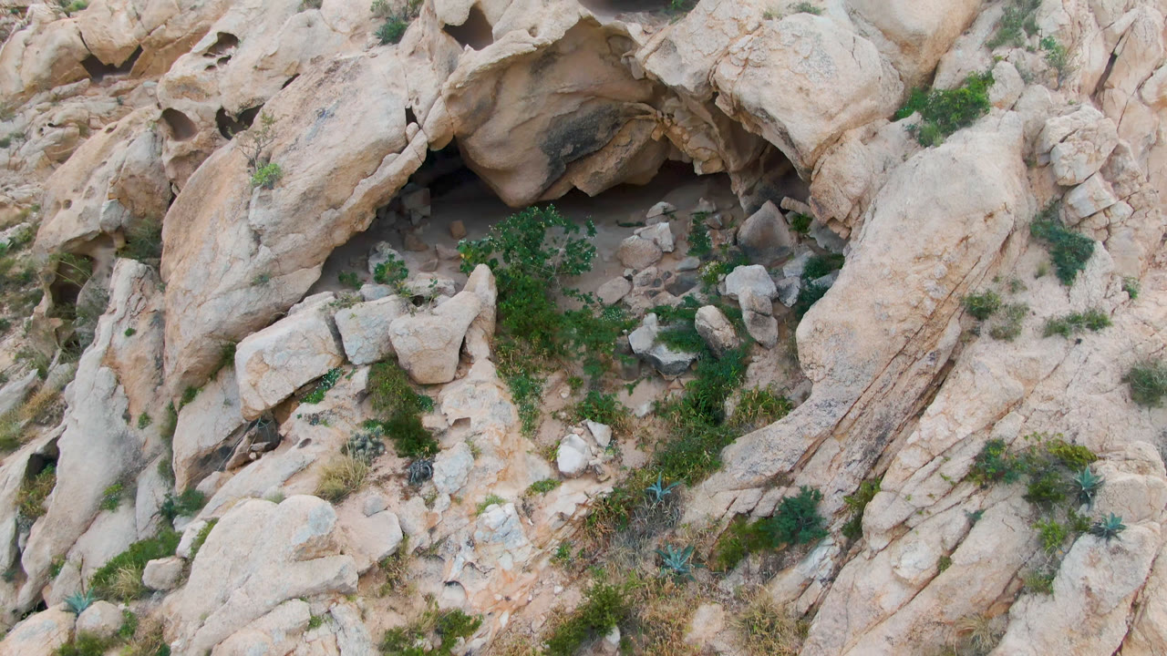 Aerial frame from Cabo Pulmo shows the cave opening near the upper rock face, scrubby green plants, loose stones, and a rough stone enclosure with no visible people.