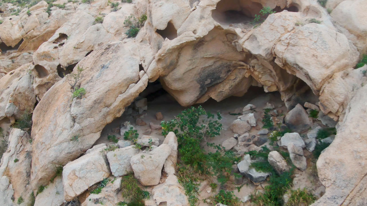 Overhead frame in Cabo Pulmo, Baja California Sur showing pale weathered rock overhangs, sandy ground, scattered stones, green brush, and no visible people.