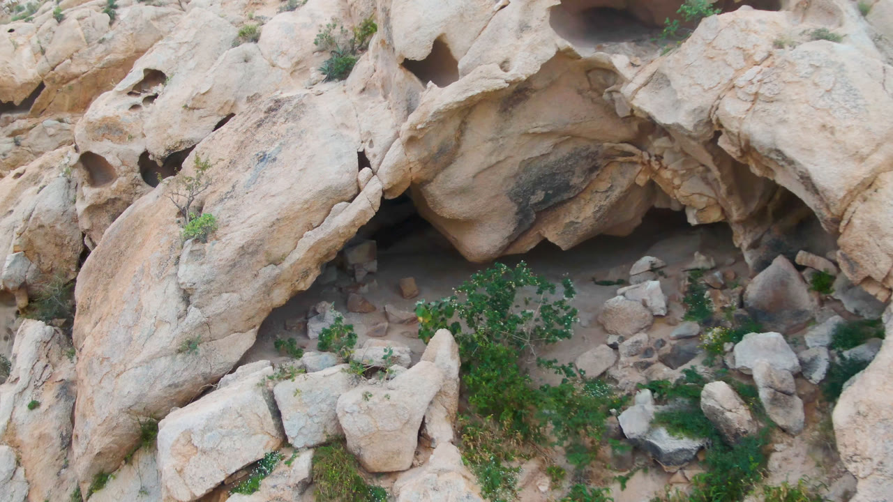 Aerial-looking frame in Cabo Pulmo, Baja California Sur with rounded tan boulders, a shaded recess beneath an overhang, small rocks, green shrubs, and no visible people.