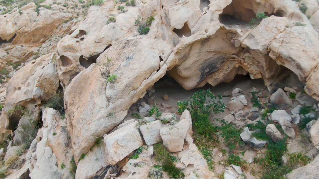 Aerial frame over Cabo Pulmo rock terrain showing pale eroded stone, a central cave-like recess, scattered boulders, green scrub, and no visible people.