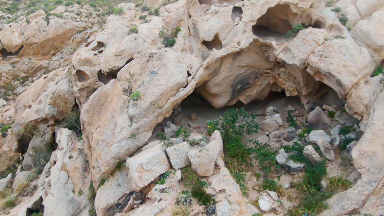 Aerial frame in Cabo Pulmo with the dark cave recess centered beneath rounded tan stone, loose rocks on sandy ground, green vegetation, and no visible people.