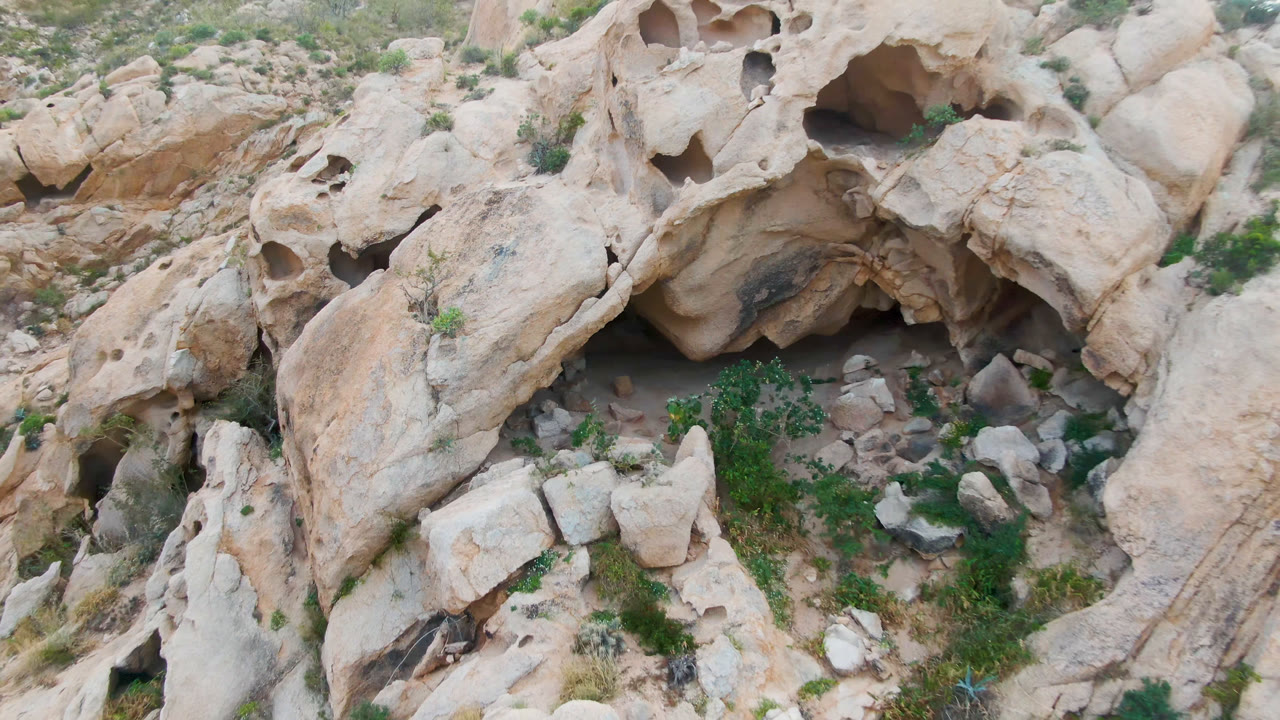 Top-down rocky terrain with honeycombed stone, cave shadow, boulders, and shrubs.