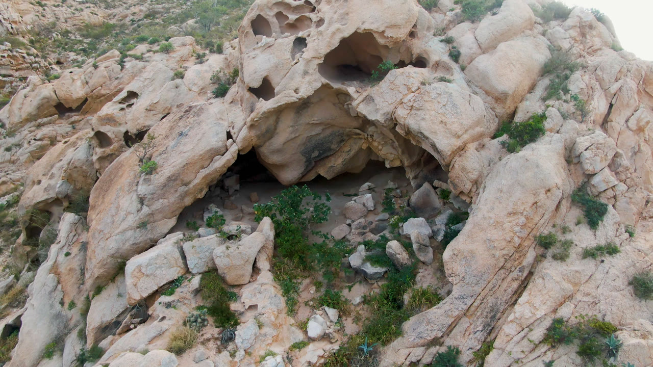 Overhead view in Cabo Pulmo, Baja California Sur, Mexico, showing pale eroded rock, dark cave recesses, scattered shrubs, and no visible people at the start of the clip.