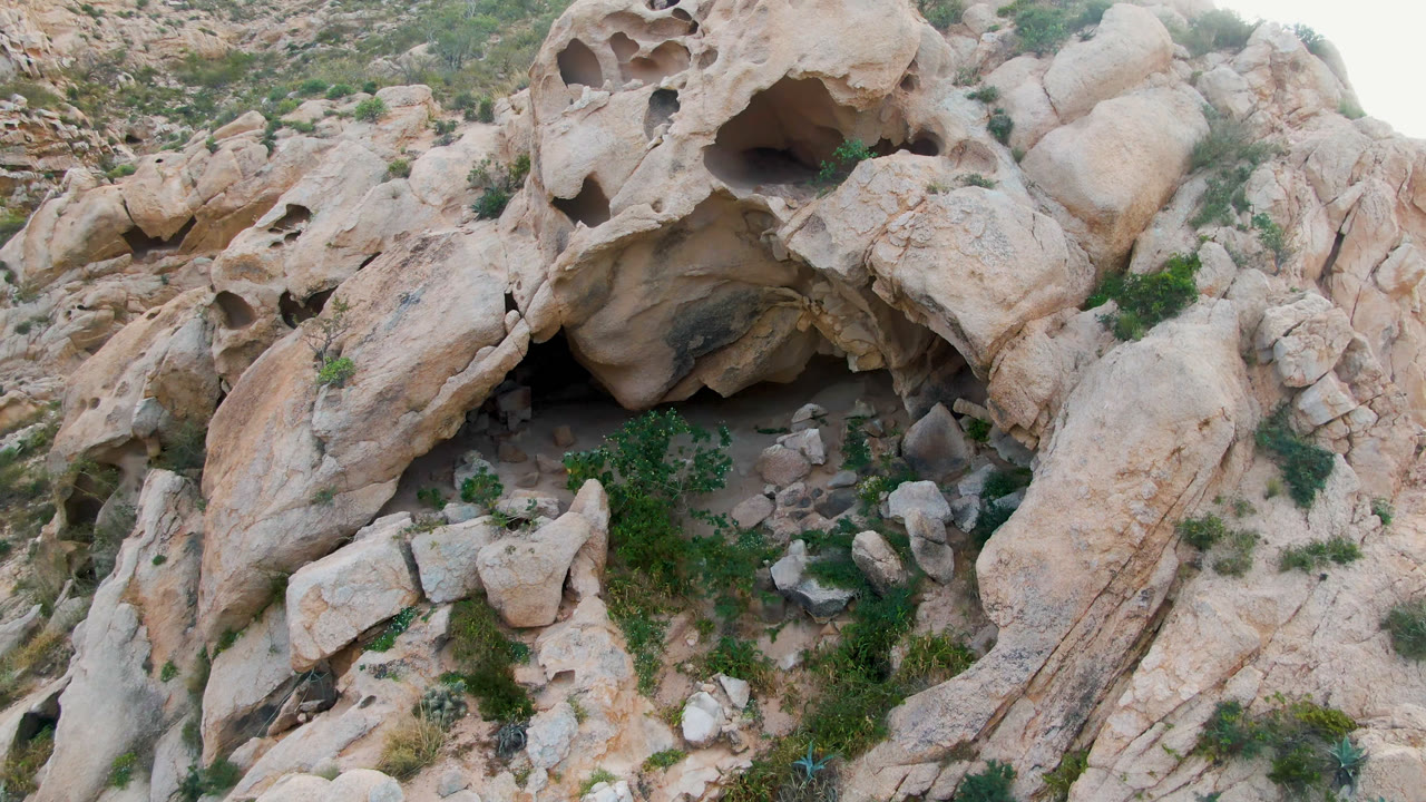 Aerial frame in Cabo Pulmo, Baja California Sur, Mexico, with the shadowed cave recess slightly more central among pale rock slabs, dry brush, and no visible people.