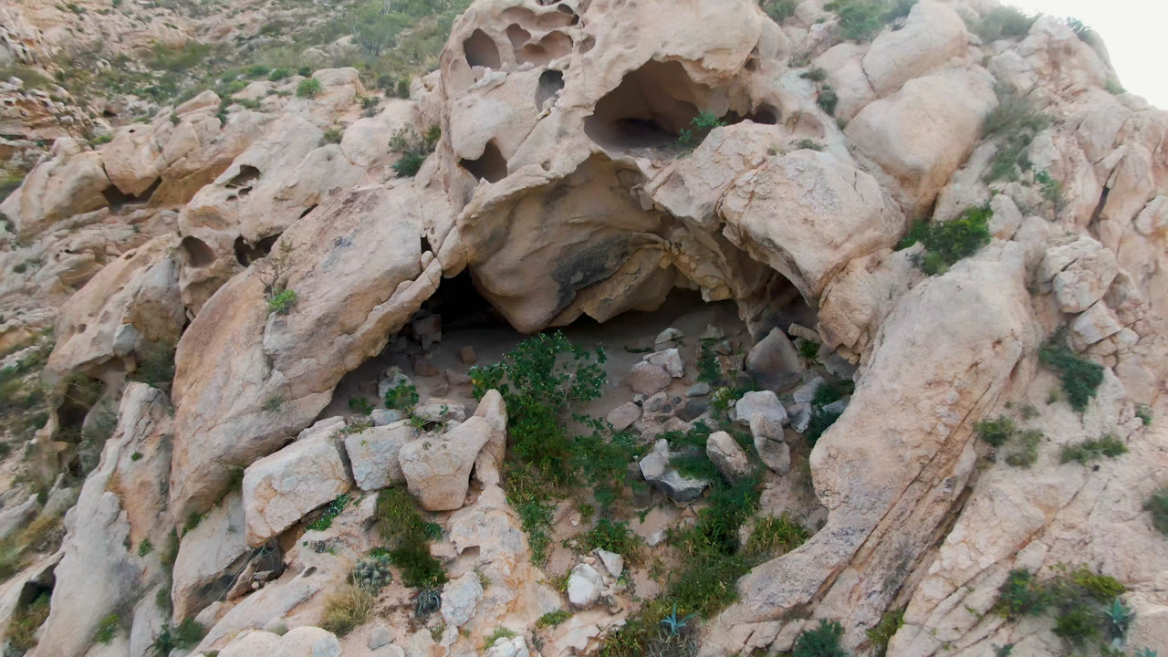 Top-down rocky slope with a cave mouth, shrubs, and broken boulders.