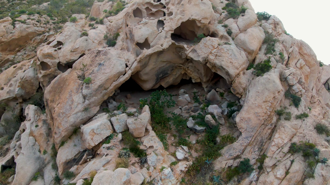 At 0:01, the cave recesses sit near the upper rock wall while loose boulders and green brush fill the lower foreground, with no visible people in Cabo Pulmo, Baja California Sur.