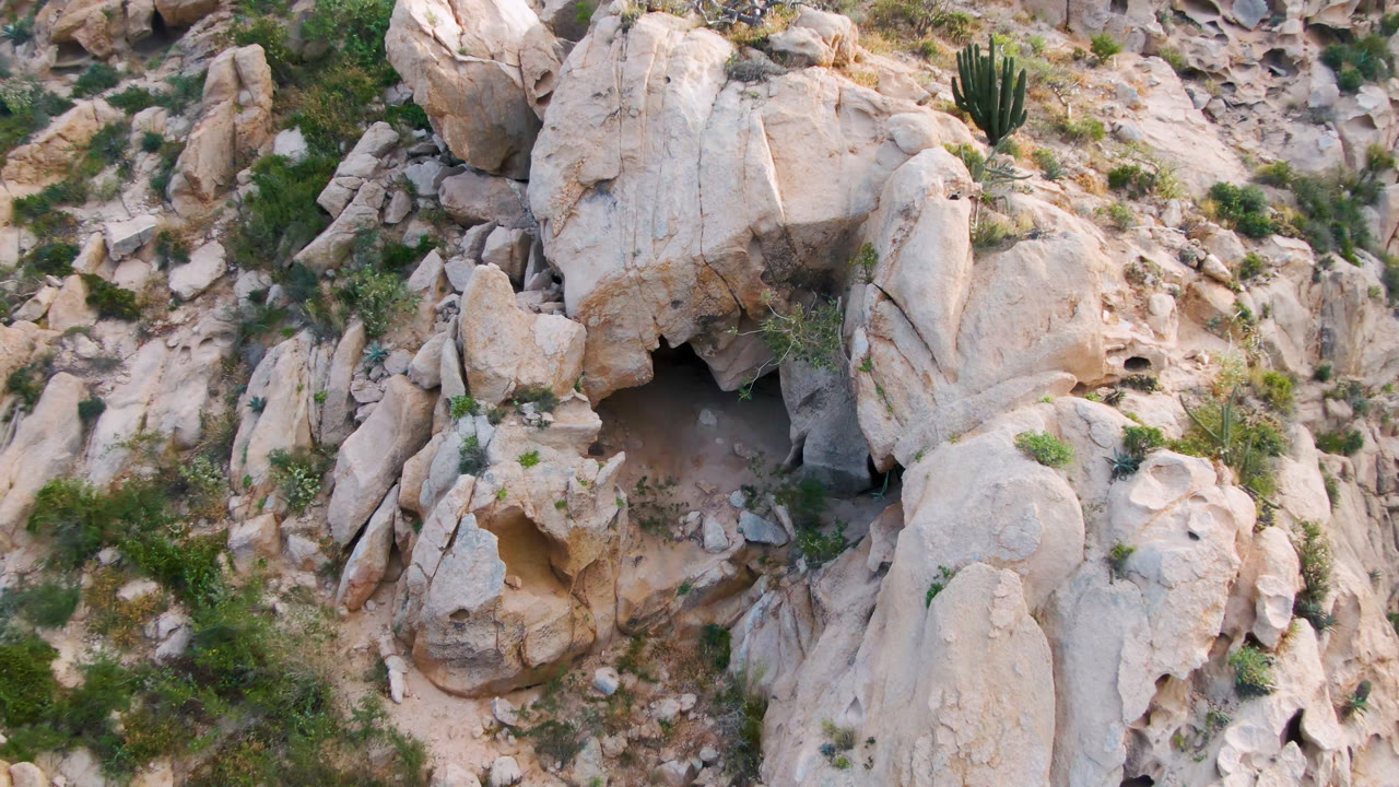 Overhead frame from Cabo Pulmo, Baja California Sur, showing pale boulders, sparse green shrubs, dry grasses, and a dark cave opening with no visible people.