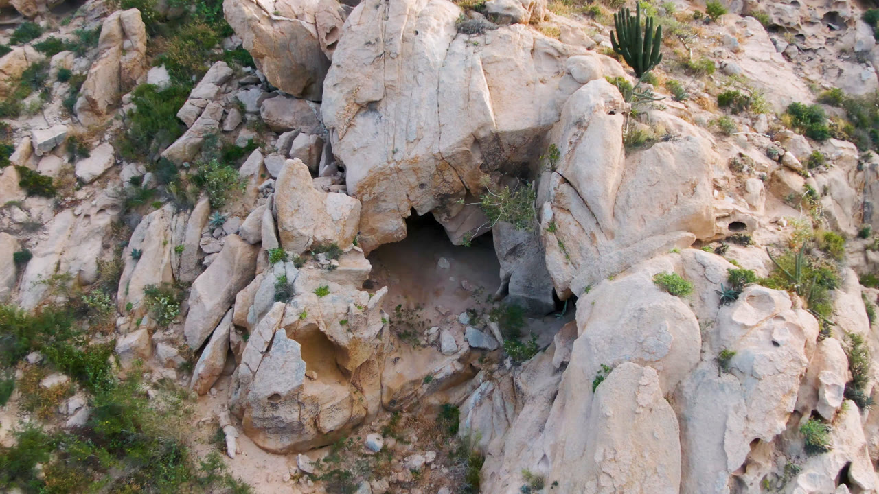 Aerial view in Cabo Pulmo, Baja California Sur, with the cave mouth centered among tan rock faces, loose stones, shrubs, grasses, and no visible people.