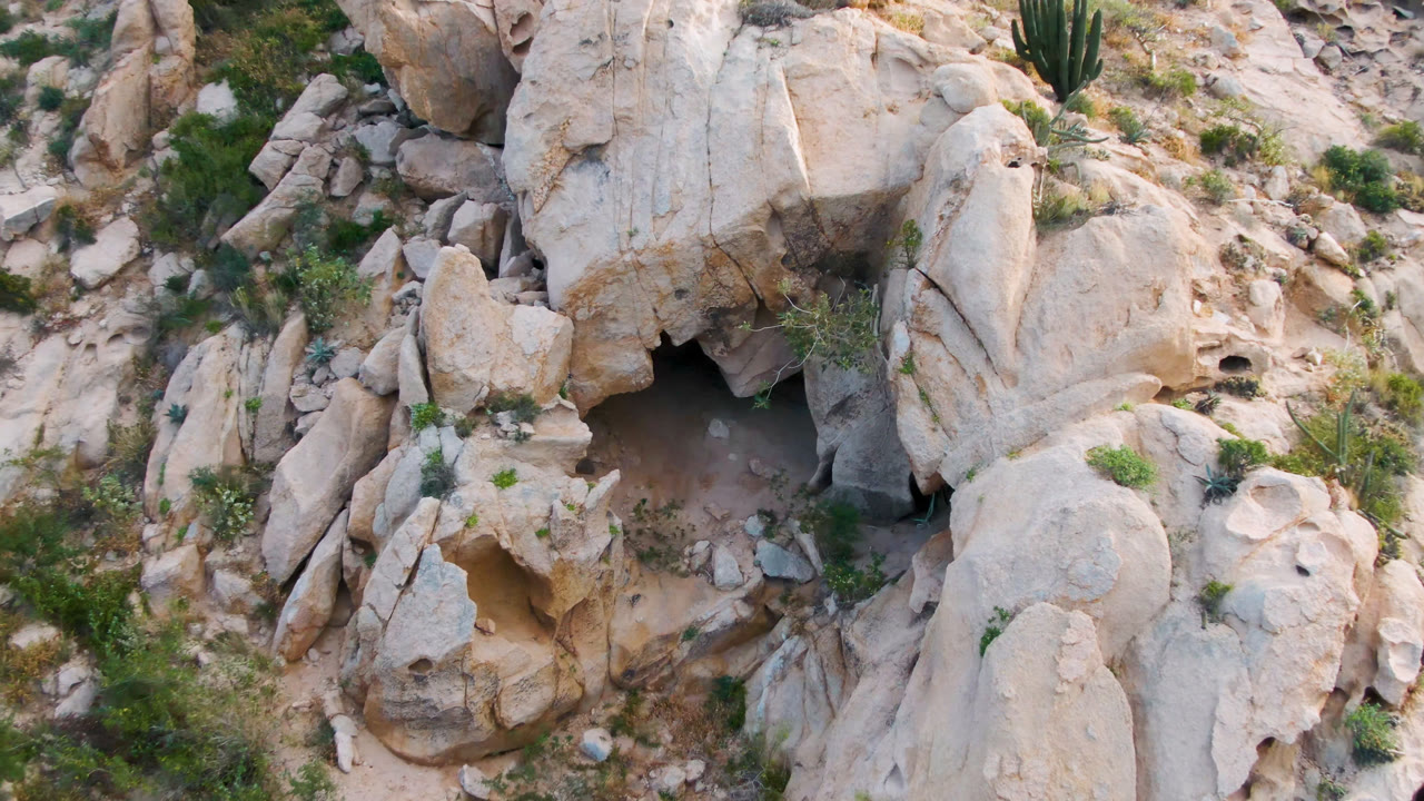 Aerial view of tan boulders, scrub plants, and a small cave opening.