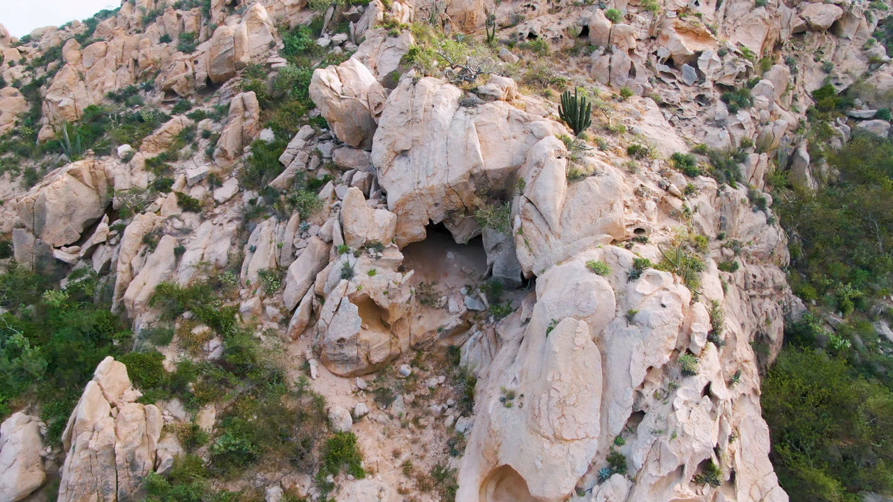 A second Cabo Pulmo aerial view holds on the same pale boulder cluster, with the cave opening lower left of center, scrub in the ravine, a tall cactus near the top, and no visible people.